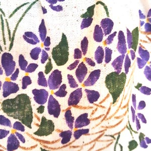 May include: White fabric with a purple and green floral pattern. The flowers are stylized and resemble violets. The fabric has a textured surface.