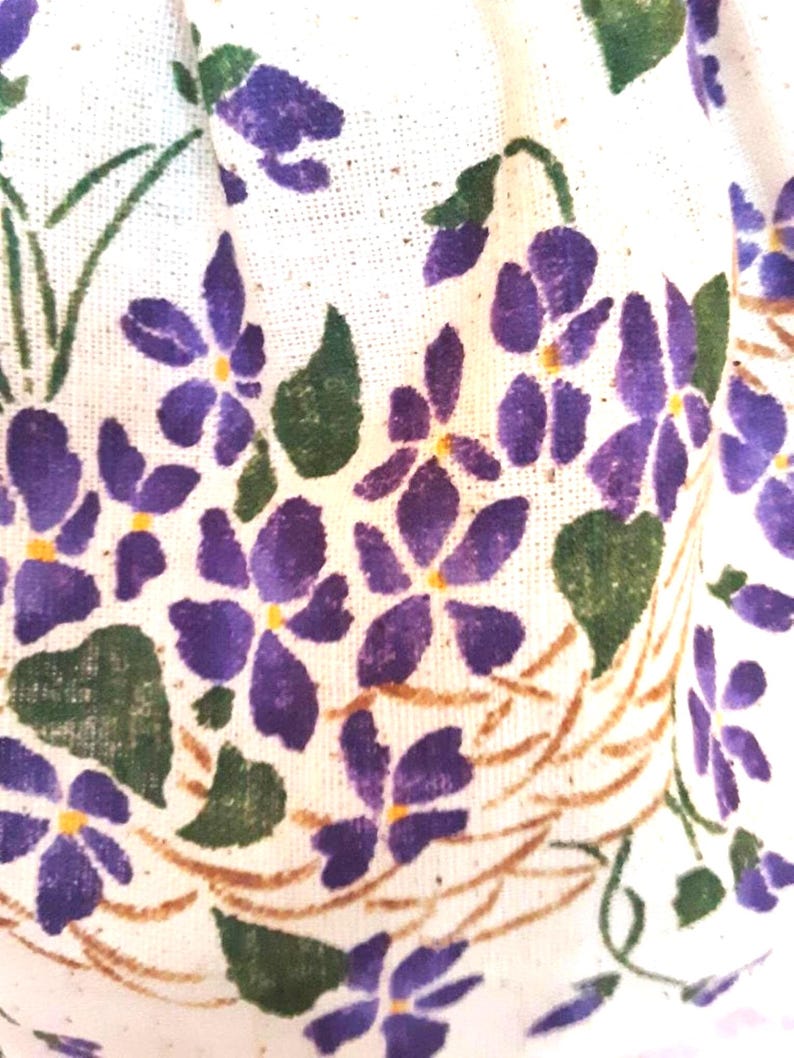 May include: White fabric with a purple and green floral pattern. The flowers are stylized and resemble violets. The fabric has a textured surface.