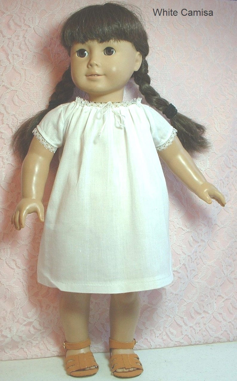 May include: A white doll wearing a white camisole with lace trim and brown sandals. The doll has long brown hair in braids and brown eyes. The doll is standing on a pink background.