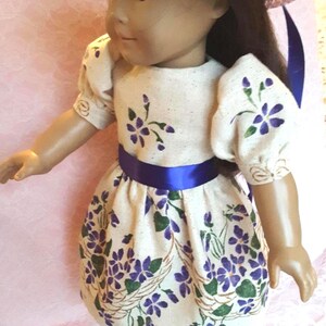 May include: A doll wearing a white dress with purple flowers and a blue sash. The doll is wearing a blue hat and blue shoes.