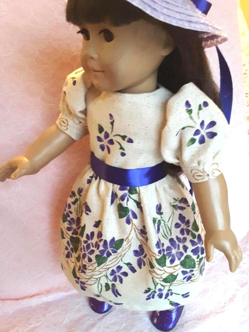 May include: A doll wearing a white dress with purple flowers and a blue sash. The doll is wearing a blue hat and blue shoes.
