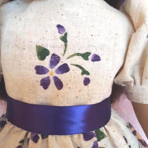 May include: A white dress with a purple satin sash and a purple floral pattern. The dress has a small purple flower with a yellow center on the bodice.