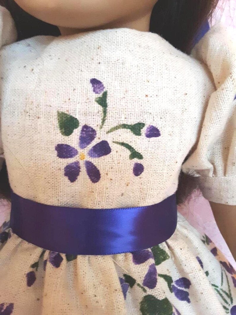 May include: A white dress with a purple satin sash and a purple floral pattern. The dress has a small purple flower with a yellow center on the bodice.