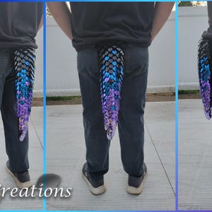 May include: A multi-colored dragon tail accessory, featuring a gradient of black, blue, and purple scales. The tail is attached to the back of a person wearing blue jeans. The tail is a unique costume piece.
