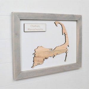 May include: A framed wooden map of Cape Cod, Massachusetts, with the town of Chatham highlighted. The map is in a gray frame and has a white background.
