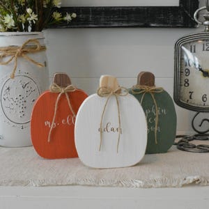 May include: Three wooden pumpkin-shaped decorations in orange, white, and green. Each pumpkin features a brown stem, a tied jute twine bow, and white cursive text. The orange pumpkin reads ms. ell, the white pumpkin reads adam, and the green pumpkin reads pumpkin.