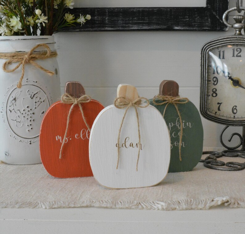 May include: Three wooden pumpkin-shaped decorations in orange, white, and green. Each pumpkin features a brown stem, a tied jute twine bow, and white cursive text. The orange pumpkin reads ms. ell, the white pumpkin reads adam, and the green pumpkin reads pumpkin.