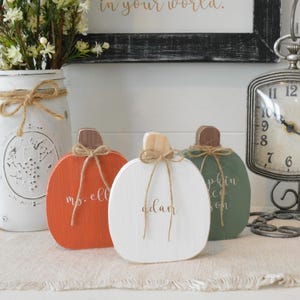 May include: Three flat wooden pumpkin decorations in orange, white, and sage green. Each pumpkin features a natural wood stem, a tied jute twine bow, and custom white script lettering. The orange pumpkin reads "ms. ell", the white one "adam", and the green one "pumpkin spice season".