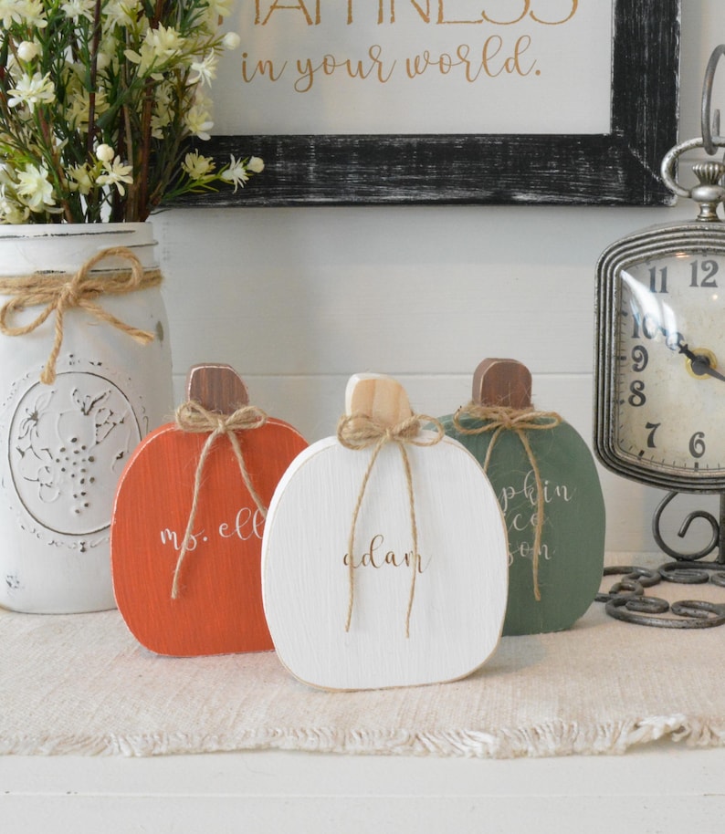 May include: Three flat wooden pumpkin decorations in orange, white, and sage green. Each pumpkin features a natural wood stem, a tied jute twine bow, and custom white script lettering. The orange pumpkin reads "ms. ell", the white one "adam", and the green one "pumpkin spice season".