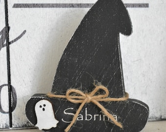 May include: A black wooden witch hat cutout with a distressed finish, featuring a small white ghost applique on the left side, the name Sabrina printed in white script, and a natural jute twine bow tied around the base of the hat crown.