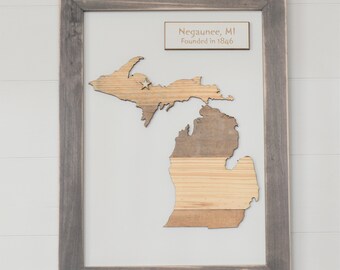 May include: A framed wall art piece featuring a wooden cutout of the state of Michigan with a small star marking Negaunee. A rectangular plaque above the map displays the text Negaunee, MI Founded in 1846. The frame is a weathered gray wood.