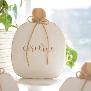 May include: A white wooden pumpkin-shaped decoration featuring a natural wood stem and a tied jute twine bow. The name "caroline" is printed in a brown script font on the center of the white surface. The object has a visible wood grain texture.