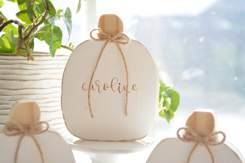 May include: A white wooden pumpkin-shaped decoration featuring a natural wood stem and a tied jute twine bow. The name "caroline" is printed in a brown script font on the center of the white surface. The object has a visible wood grain texture.