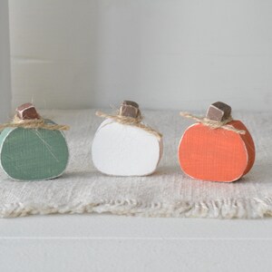 May include: Three small decorative pumpkins crafted from wood, featuring a sage green, a white, and an orange pumpkin. Each pumpkin is topped with a small brown wooden stem and a piece of natural jute twine tied in a simple bow around the base.