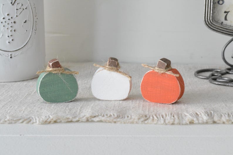 May include: Three small decorative pumpkins crafted from wood, featuring a sage green, a white, and an orange pumpkin. Each pumpkin is topped with a small brown wooden stem and a piece of natural jute twine tied in a simple bow around the base.