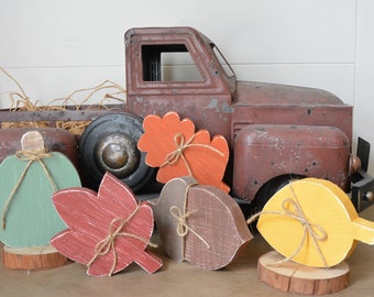 May include: A rustic metal truck in a weathered brown finish sits behind several wooden cutouts including a sage green pumpkin, a red maple leaf, a brown acorn, an orange oak leaf, and a yellow leaf, each tied with a thin jute twine bow.