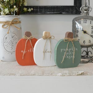 May include: Three flat wooden pumpkin decorations in orange, white, and sage green. Each pumpkin features a brown wooden stem, a tied jute twine bow, and white cursive text. The orange pumpkin reads ms. ell, the white reads adam, and the green reads pumpkin spice season.