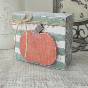 May include: A rectangular wooden block sign featuring horizontal green and white distressed stripes. A flat, orange wooden pumpkin cutout with a brown stem is attached to the front, wrapped with a thin, natural-colored jute twine bow.
