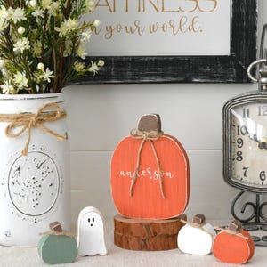May include: A flat, orange wooden pumpkin cutout with the name "anderson" in white script, tied with a jute twine bow at the brown stem. It sits on a round wood slice, surrounded by a small white ghost, a green pumpkin, and two smaller pumpkins.