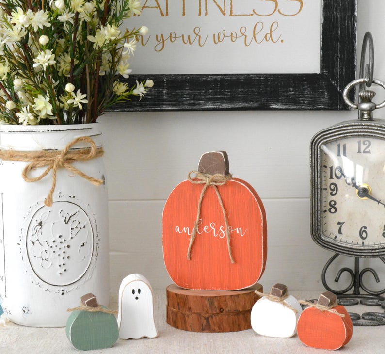 May include: A flat, orange wooden pumpkin cutout with the name "anderson" in white script, tied with a jute twine bow at the brown stem. It sits on a round wood slice, surrounded by a small white ghost, a green pumpkin, and two smaller pumpkins.