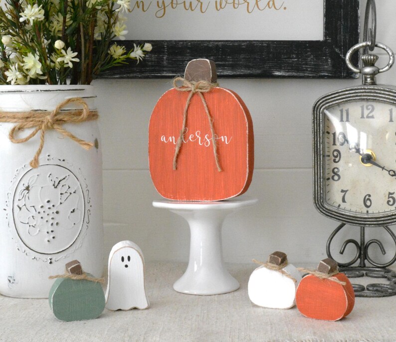 May include: A flat, orange wooden pumpkin cutout featuring the name "anderson" in white script, topped with a brown stem and a tied jute twine bow, displayed on a white pedestal stand alongside smaller pumpkin and ghost-shaped wooden figures.
