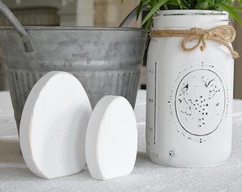 May include: Two white wooden eggs, one larger than the other, sit on a white tablecloth. A galvanized metal bucket and a white painted mason jar with a black grape design are in the background.