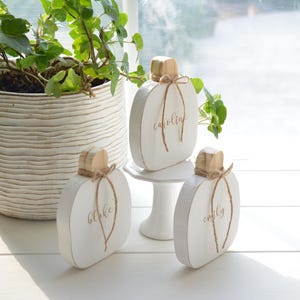 May include: Three white wooden pumpkin-shaped decorations with natural wood stems and twine bows. Each pumpkin features a name written in gold script: Blake, Caroline, and Emily. The pumpkins are displayed on a white surface with one elevated on a small white pedestal.