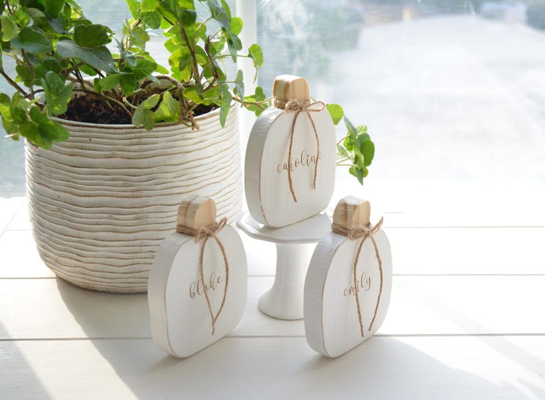May include: Three white wooden pumpkin-shaped decorations with natural wood stems and twine bows. Each pumpkin features a name written in gold script: Blake, Caroline, and Emily. The pumpkins are displayed on a white surface with one elevated on a small white pedestal.