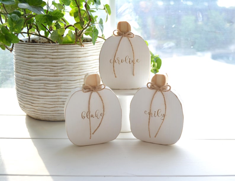 May include: Three white wooden pumpkin-shaped decorations with natural wood stems and jute twine bows. Each pumpkin features a name written in cursive script: Caroline on the center pumpkin, Blake on the left, and Emily on the right side.
