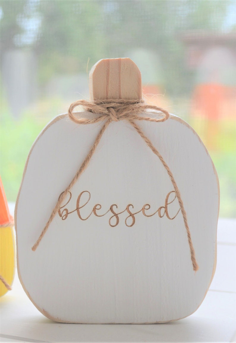 May include: A white wooden pumpkin-shaped decoration featuring the word blessed in brown cursive script. The pumpkin has a natural light wood stem and is adorned with a thin brown twine bow tied around the base of the stem, resting on a surface.