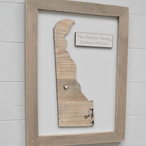 May include: A wooden cutout of the state of Delaware in a light wood frame. The cutout is made of reclaimed wood and has a small white heart in the center. A small wooden sign with the text "The Fletcher Family Sunflower Hill Farm" is attached to the frame.