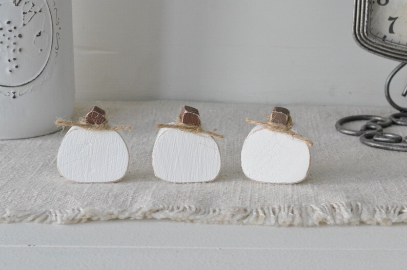 May include: Three small, white-painted wooden pumpkin cutouts arranged in a row. Each pumpkin features a brown wooden stem and a thin piece of natural jute twine tied around the base of the stem, creating a simple, rustic decorative accent.