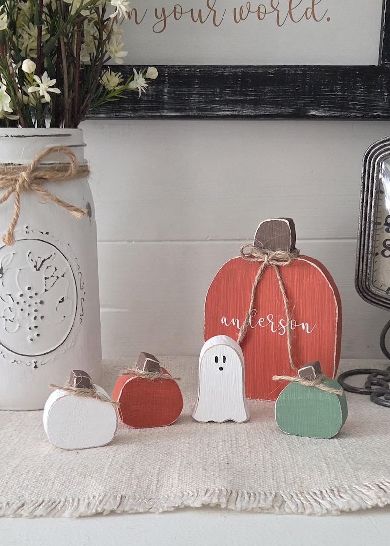 May include: A set of decorative wooden figures including three pumpkins in white, orange, and green, and one white ghost. The largest orange pumpkin features the name "anderson" in white script and a brown stem, with all items tied with twine.