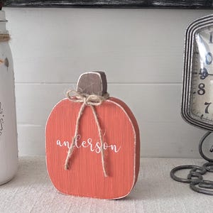 May include: A flat, orange wooden pumpkin cutout with a brown stem and a jute twine bow tied around the top. The word "anderson" is written in white cursive script across the center of the pumpkin. The surface shows a visible wood grain texture.