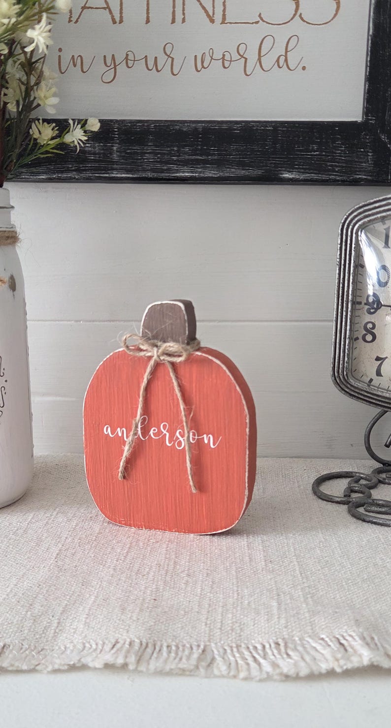 May include: A flat, orange wooden pumpkin cutout with a brown stem and a jute twine bow tied around the top. The word "anderson" is written in white cursive script across the center of the pumpkin. The surface shows a visible wood grain texture.