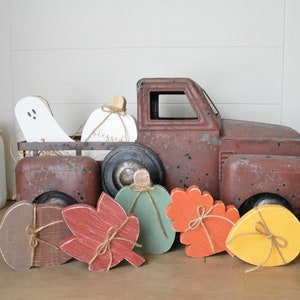 May include: A rustic brown metal toy truck carrying a white wooden ghost and a white pumpkin with the word blessed written on it. In front of the truck are five wooden cutouts shaped like an acorn, a red leaf, a green pumpkin, an orange leaf, and a yellow leaf.