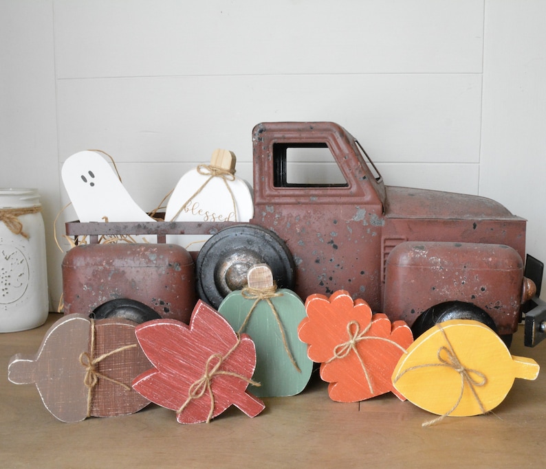 May include: A rustic brown metal toy truck carrying a white wooden ghost and a white pumpkin with the word blessed written on it. In front of the truck are five wooden cutouts shaped like an acorn, a red leaf, a green pumpkin, an orange leaf, and a yellow leaf.