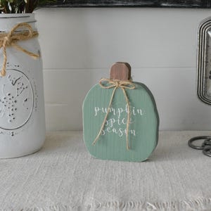 May include: A sage green wooden pumpkin cutout featuring a brown stem and a jute twine bow tied around the top. The words "pumpkin spice season" are printed in white cursive script on the front of the pumpkin, which has a visible wood grain texture.