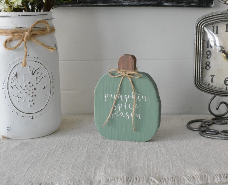 May include: A sage green wooden pumpkin cutout featuring a brown stem and a jute twine bow tied around the top. The words "pumpkin spice season" are printed in white cursive script on the front of the pumpkin, which has a visible wood grain texture.