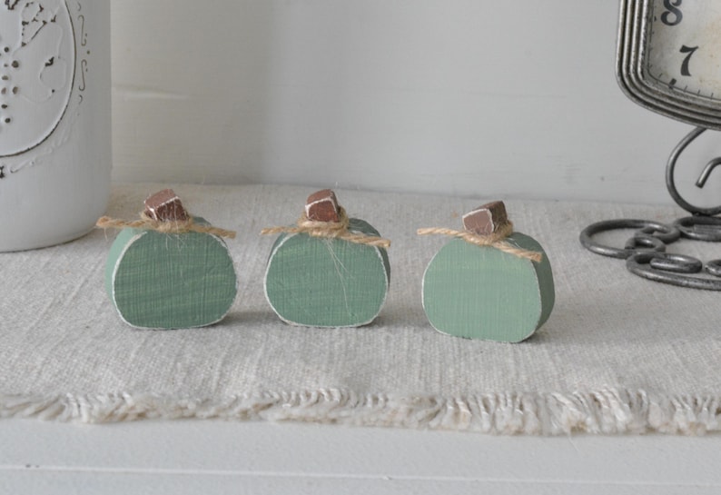 May include: Three small, sage green wooden pumpkins arranged in a row. Each pumpkin features a flat, rounded shape with a distressed white edge, a small brown wooden stem, and a thin piece of natural jute twine tied around the base of the stem.