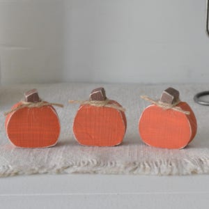 May include: Three small, flat, orange-painted wooden pumpkins arranged in a row. Each pumpkin features a dark brown wooden stem and a thin piece of natural jute twine tied in a simple bow around the base of the stem. The edges show white paint.