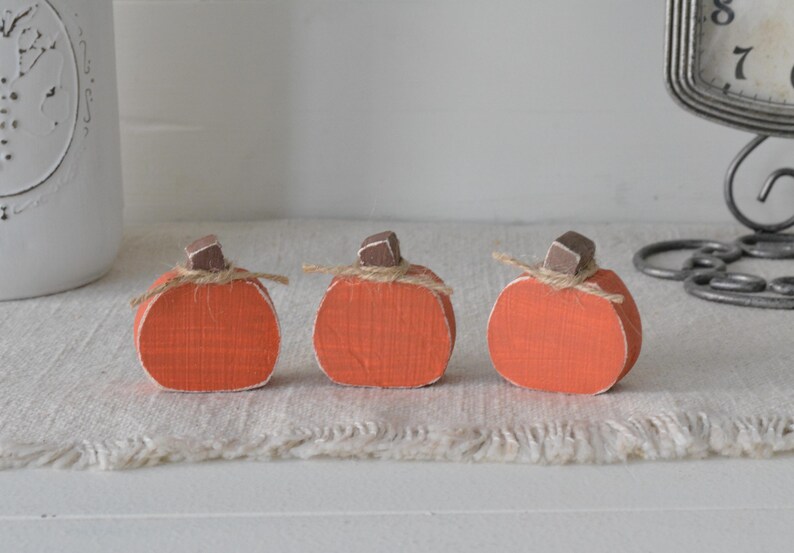 May include: Three small, flat, orange-painted wooden pumpkins arranged in a row. Each pumpkin features a dark brown wooden stem and a thin piece of natural jute twine tied in a simple bow around the base of the stem. The edges show white paint.