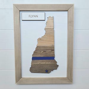 May include: Framed wooden art piece featuring the state of New Hampshire. The artwork is made of various wood tones with a blue stripe and a blue star. The name "FLYNN" is displayed above the state outline.