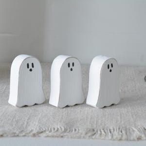 May include: Three small, white, ghost-shaped wooden figures standing in a row. Each figure features a hand-painted black face with two oval eyes and a small circular mouth. The wood has a visible grain texture with slightly distressed edges.