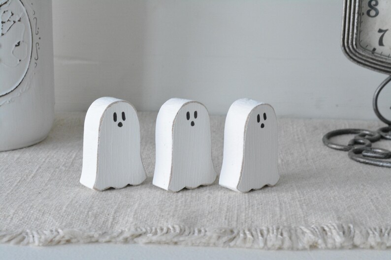 May include: Three small, white, ghost-shaped wooden figures standing in a row. Each figure features a hand-painted black face with two oval eyes and a small circular mouth. The wood has a visible grain texture with slightly distressed edges.