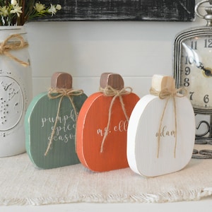 May include: Three flat wooden pumpkin decorations in green, orange, and white. Each pumpkin features a brown or natural wood stem tied with a thin jute twine bow. The green pumpkin reads pumpkin spice season, the orange reads ms. ell, and the white reads adam.