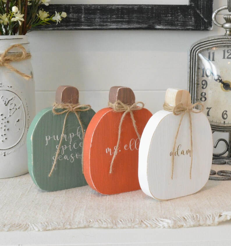 May include: Three flat wooden pumpkin decorations in green, orange, and white. Each pumpkin features a brown or natural wood stem tied with a thin jute twine bow. The green pumpkin reads pumpkin spice season, the orange reads ms. ell, and the white reads adam.