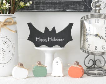 May include: A black wooden bat cutout with the white text Happy Halloween sits on a white pedestal. Below are four small wooden figures: a white pumpkin, a green pumpkin, a white ghost, and an orange pumpkin, each with a brown stem and twine.