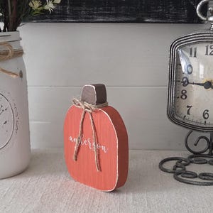 May include: A flat, orange wooden pumpkin cutout with a dark brown stem and a rustic jute twine bow tied around the top. The name "anderson" is written in white cursive script across the center of the orange surface of the wooden pumpkin.