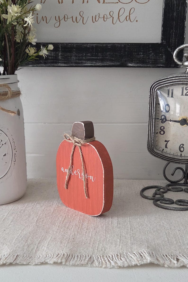 May include: A flat, orange wooden pumpkin cutout with a dark brown stem and a rustic jute twine bow tied around the top. The name "anderson" is written in white cursive script across the center of the orange surface of the wooden pumpkin.